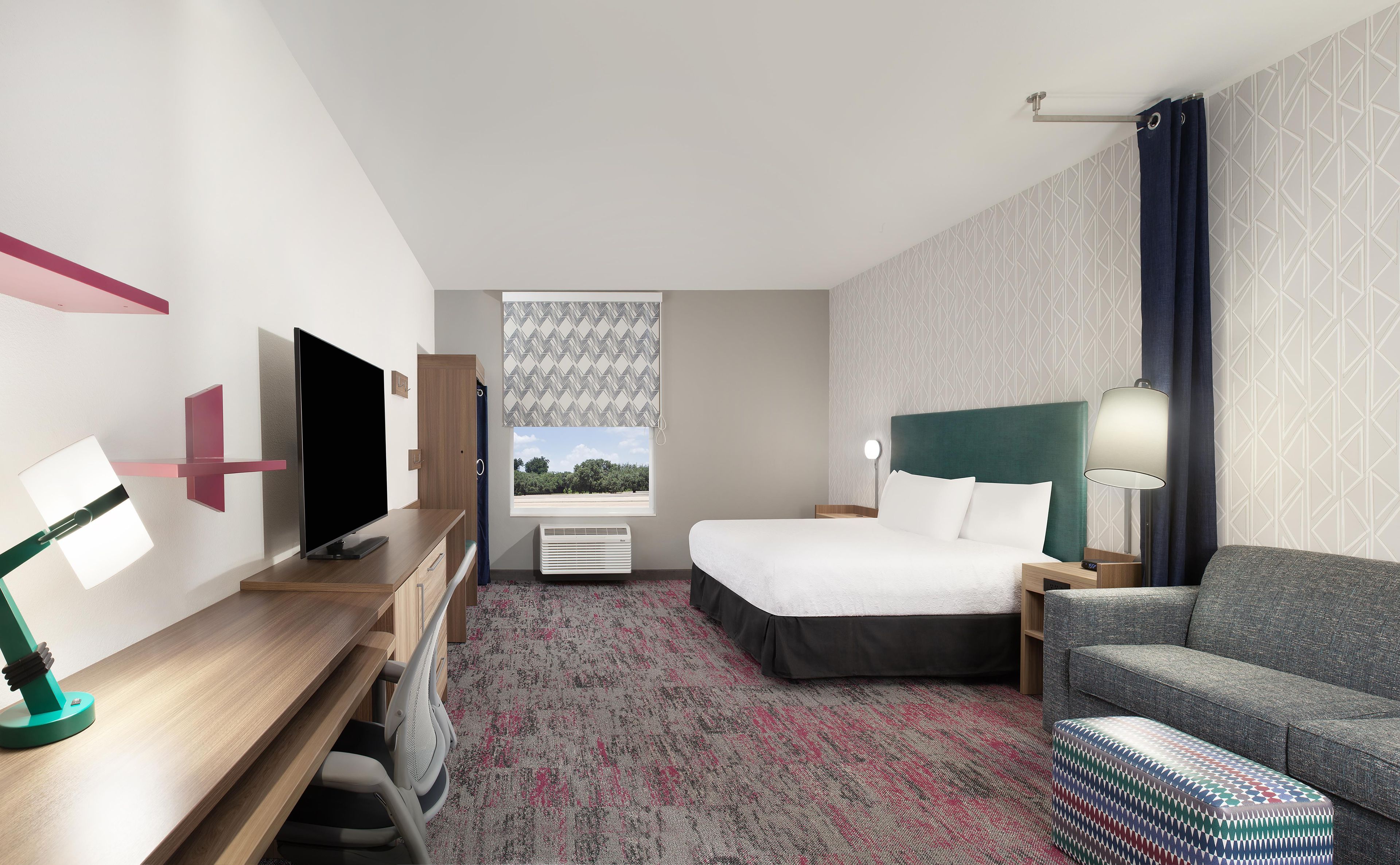 Quarto de hotel com cama, escrivaninha com televisão, sofá, tapete estampado e janela com vista.