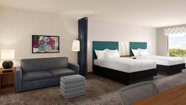 Une chambre d’hôtel avec un canapé gris, deux lits dotés de têtes de lit turquoise, un tableau abstrait aux couleurs vives, et une vue sur la verdure par la fenêtre.