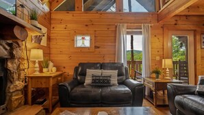 A cozy cabin interior with a fireplace, leather sofa, wooden walls, and a view of the outdoors. - Nice View Log Cabin Fireplace Hot Tub Pool Table (Sevierville)
