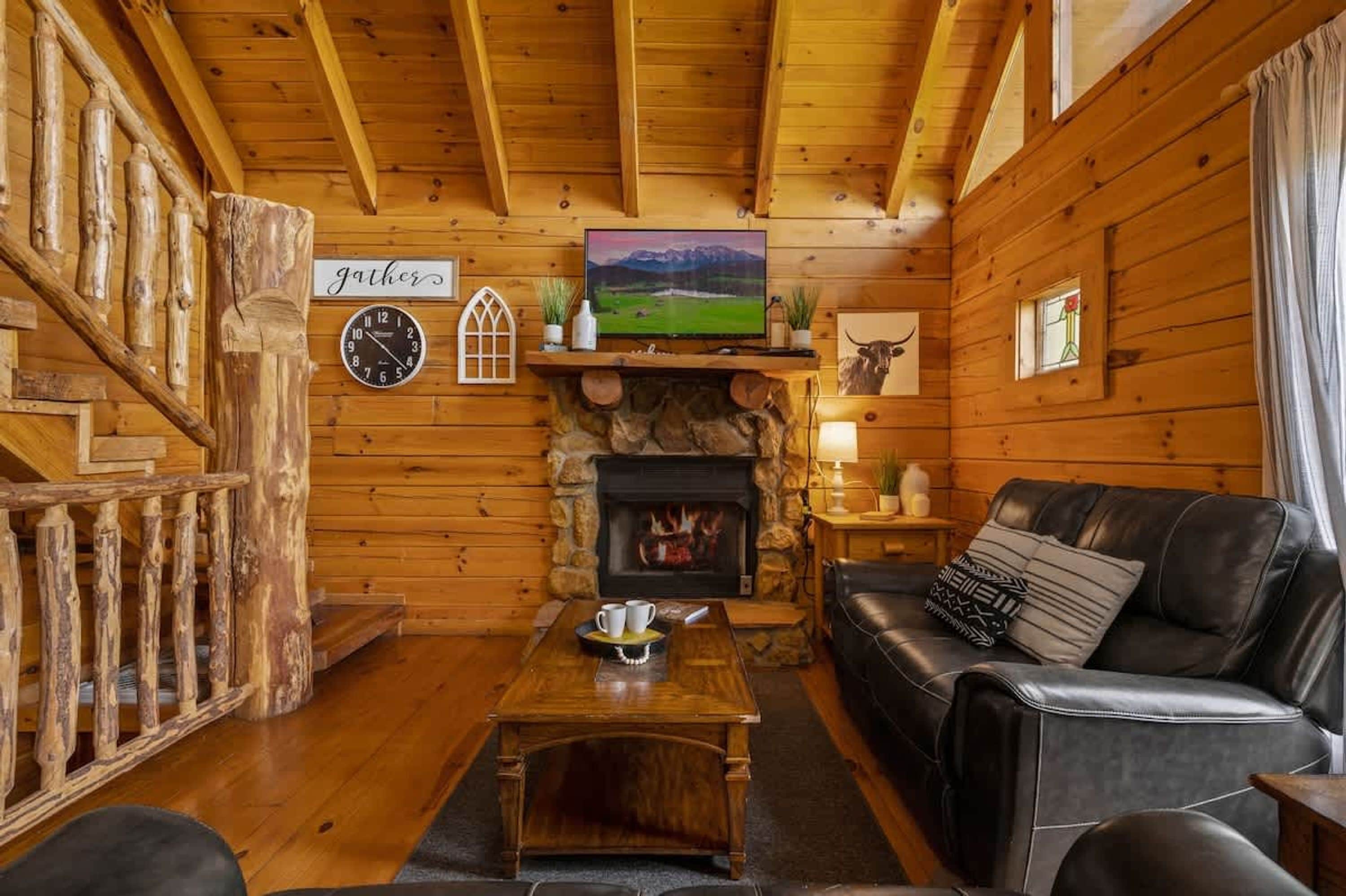 A cozy log cabin interior with a fireplace, a television mounted above it, a leather sofa, and a wooden coffee table.