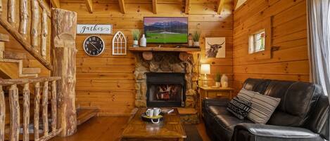 A cozy log cabin interior with a fireplace, a television mounted above it, a leather sofa, and a wooden coffee table.