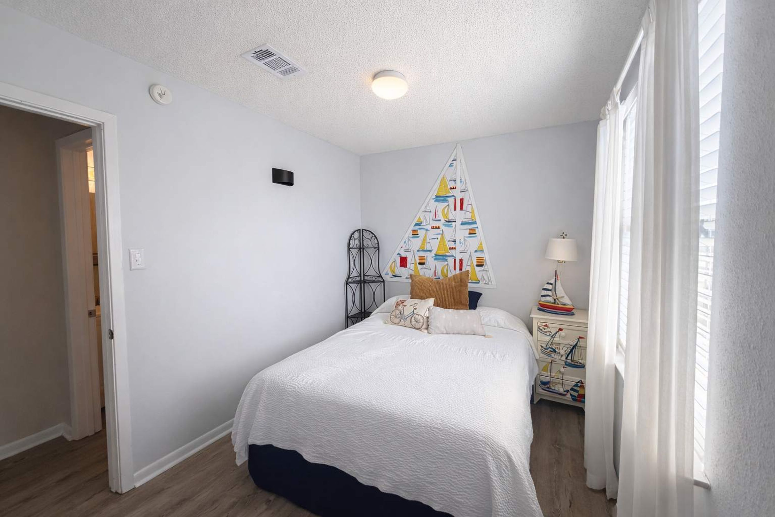 Une chambre à coucher comprenant un lit, une table de chevet, une lampe, une fenêtre avec des rideaux et un élément de décoration murale.