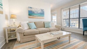 Un soggiorno con un divano color beige, un tavolino da caffè bianco, un tappeto a righe, una lampada e un quadro raffigurante una spiaggia.