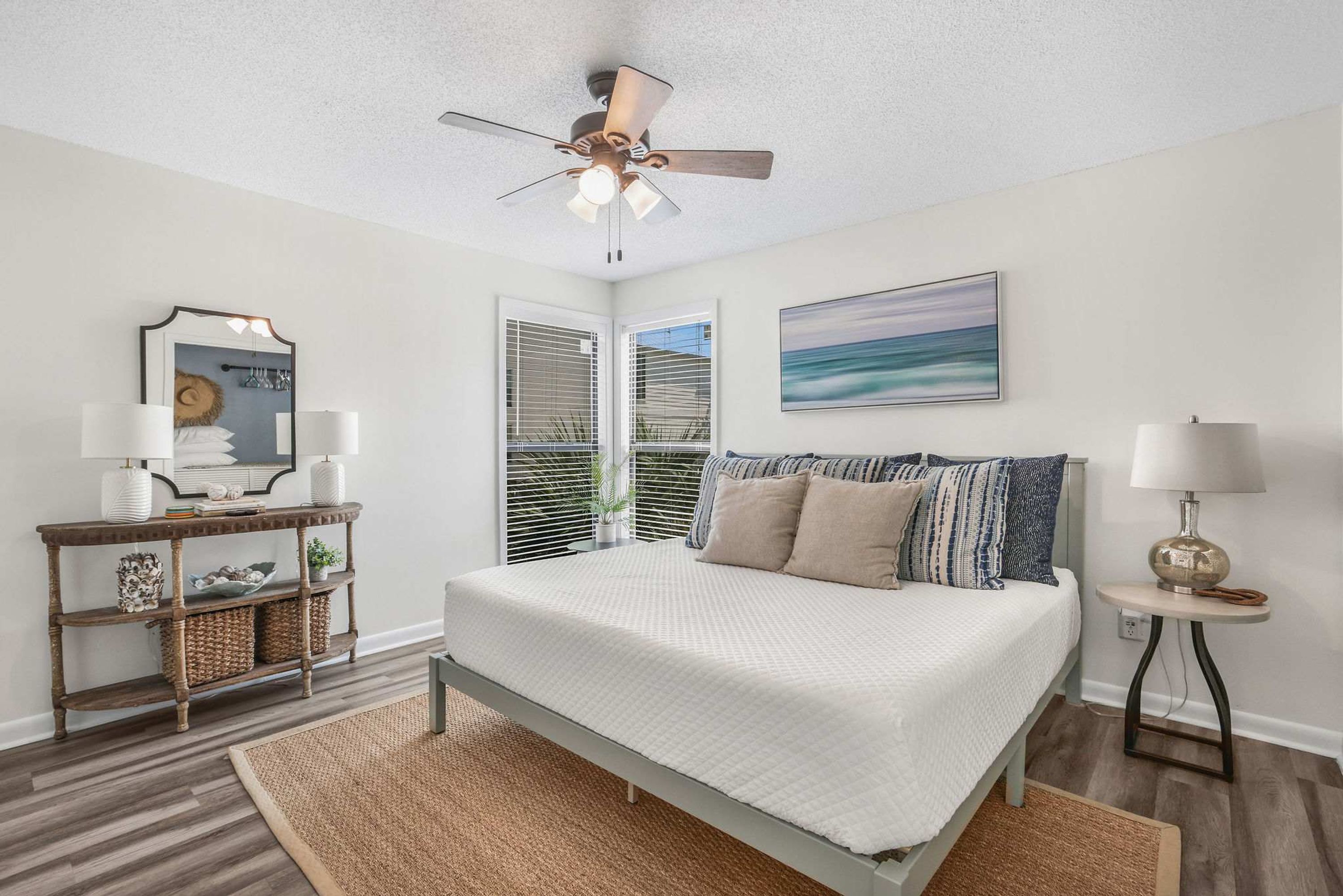 A bedroom with a bed, a ceiling fan, a console table with lamps, a wall-mounted mirror, and a painting.