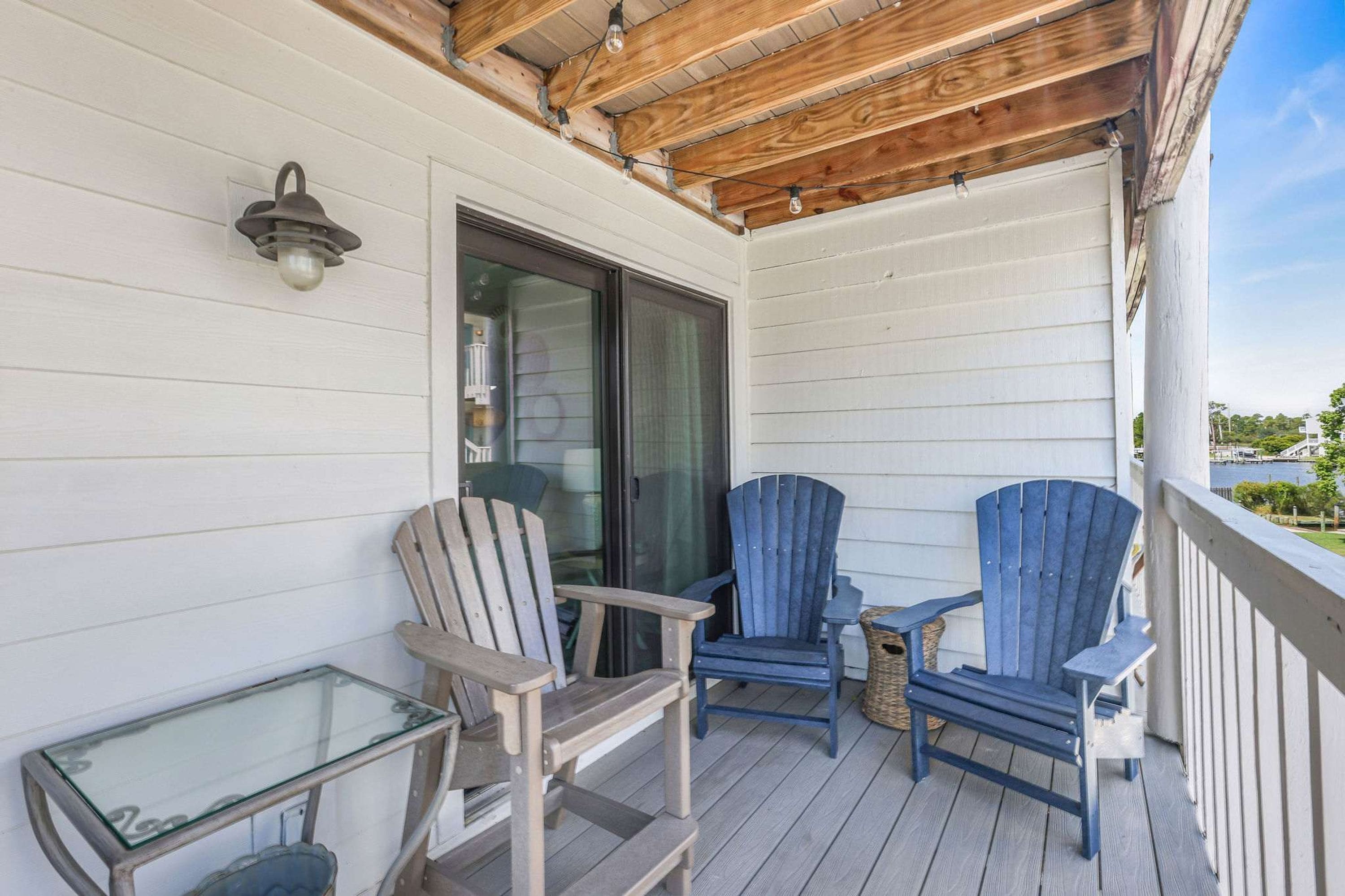 A wooden deck with two Adirondack chairs and a glass table.
