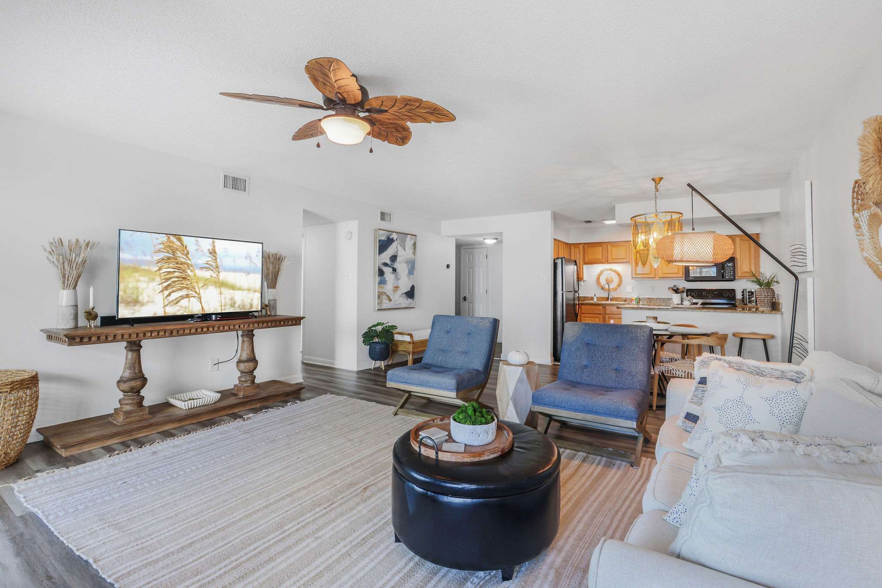 A modern living room with a flat-screen TV, a ceiling fan, and a kitchen in the background.