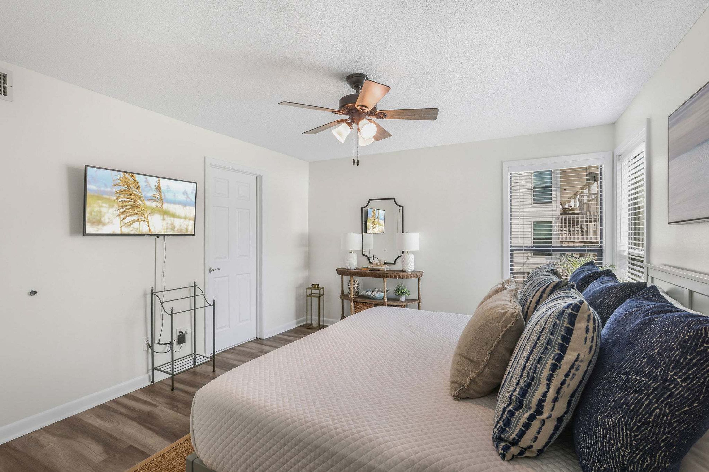 A bedroom with a bed, a ceiling fan, a window with blinds, a bedside table with a lamp, and a wall-mounted TV.