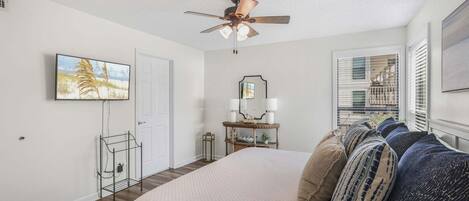 A bedroom with a bed, a ceiling fan, a window with blinds, a bedside table with a lamp, and a wall-mounted TV.