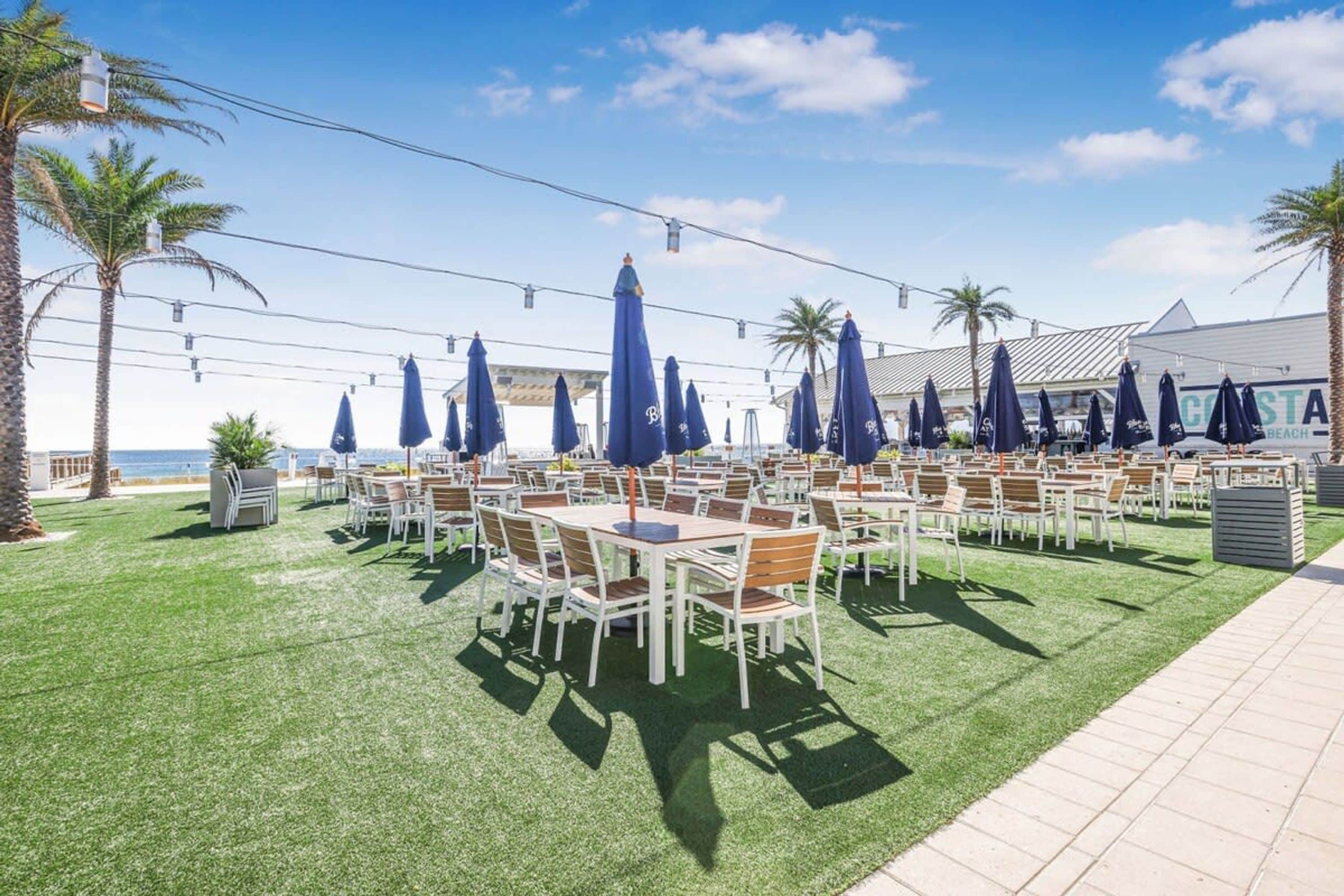 Egy szabadtéri étkezőhely, ahol asztalok, székek, napernyők és pálmafák találhatók.