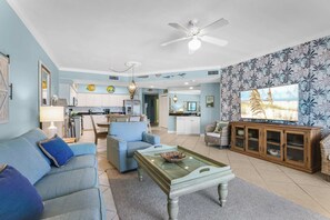 A spacious living room with a sofa, coffee table, and TV. There is a kitchen in the background with white cabinets and a dining area. - Admirals Quarters 806- Luxury Beachfront 8th fl (Orange Beach)