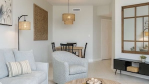 Ein modernes Wohnzimmer mit einem hellblauen Sofa, einem gemusterten Sessel, einem runden Holztisch, einem Deckenventilator und einem Essbereich im Hintergrund.