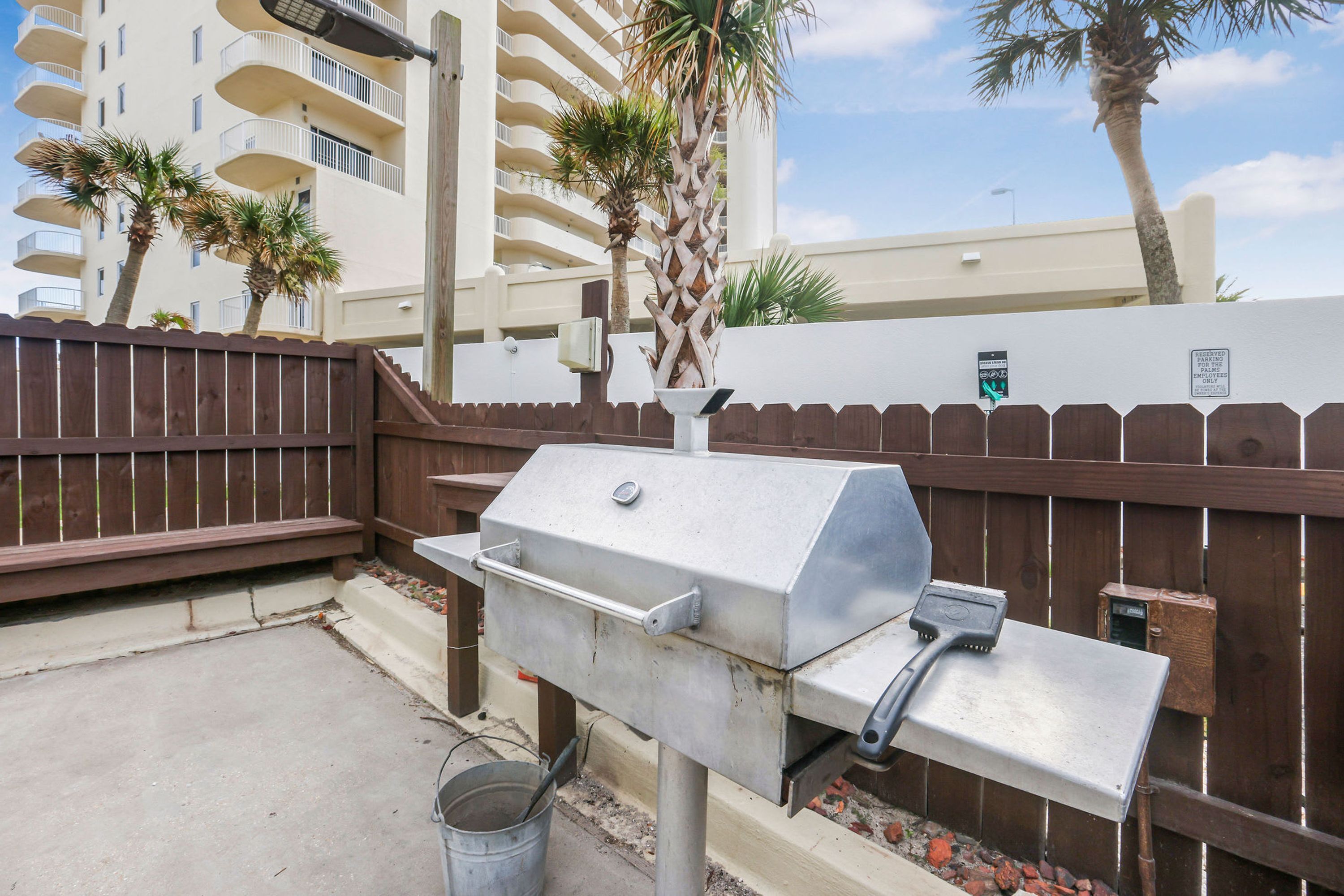 A barbecue grill with a metal grate and a side burner, positioned against a wooden fence with a palm tree in the background.