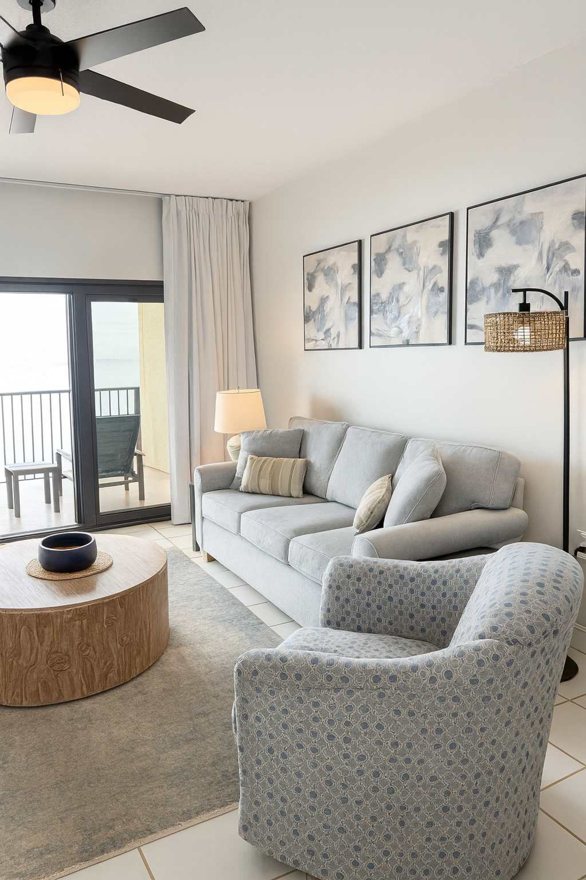 A modern living room with a grey sofa, patterned armchair, and a round wooden coffee table. There are abstract paintings on the wall and a floor lamp.