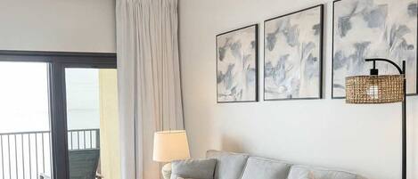 A modern living room with a grey sofa, patterned armchair, and a round wooden coffee table. There are abstract paintings on the wall and a floor lamp.
