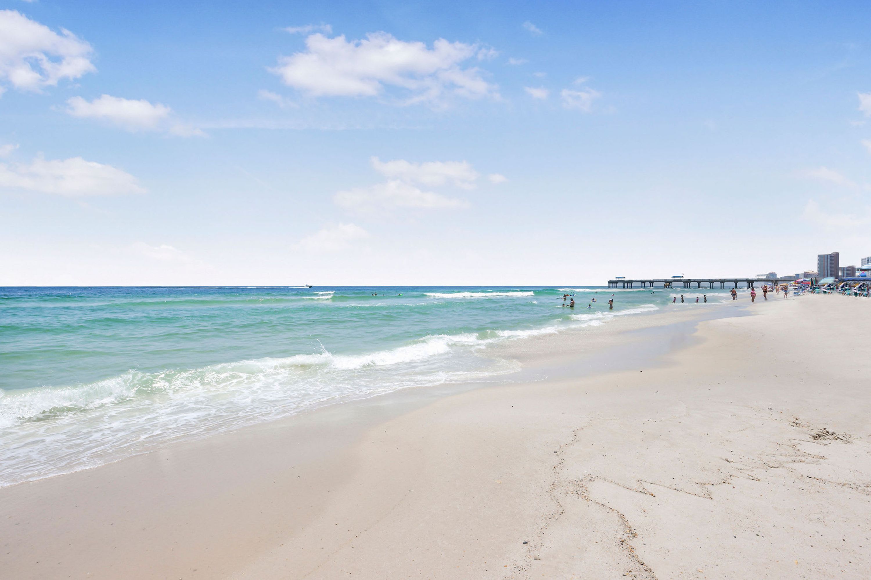 A wide sandy beach with gentle waves, a pier in the background, and a clear sky.