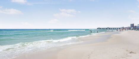 A wide sandy beach with gentle waves, a pier in the background, and a clear sky.