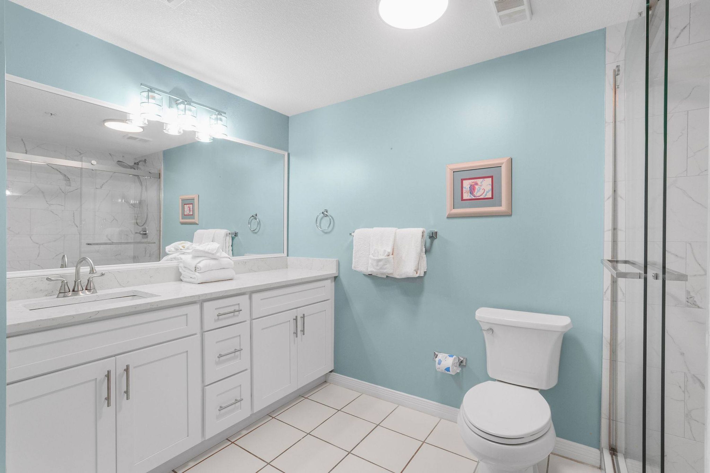 A bathroom with a white sink, mirror, and toilet, light blue walls, and a shower area.