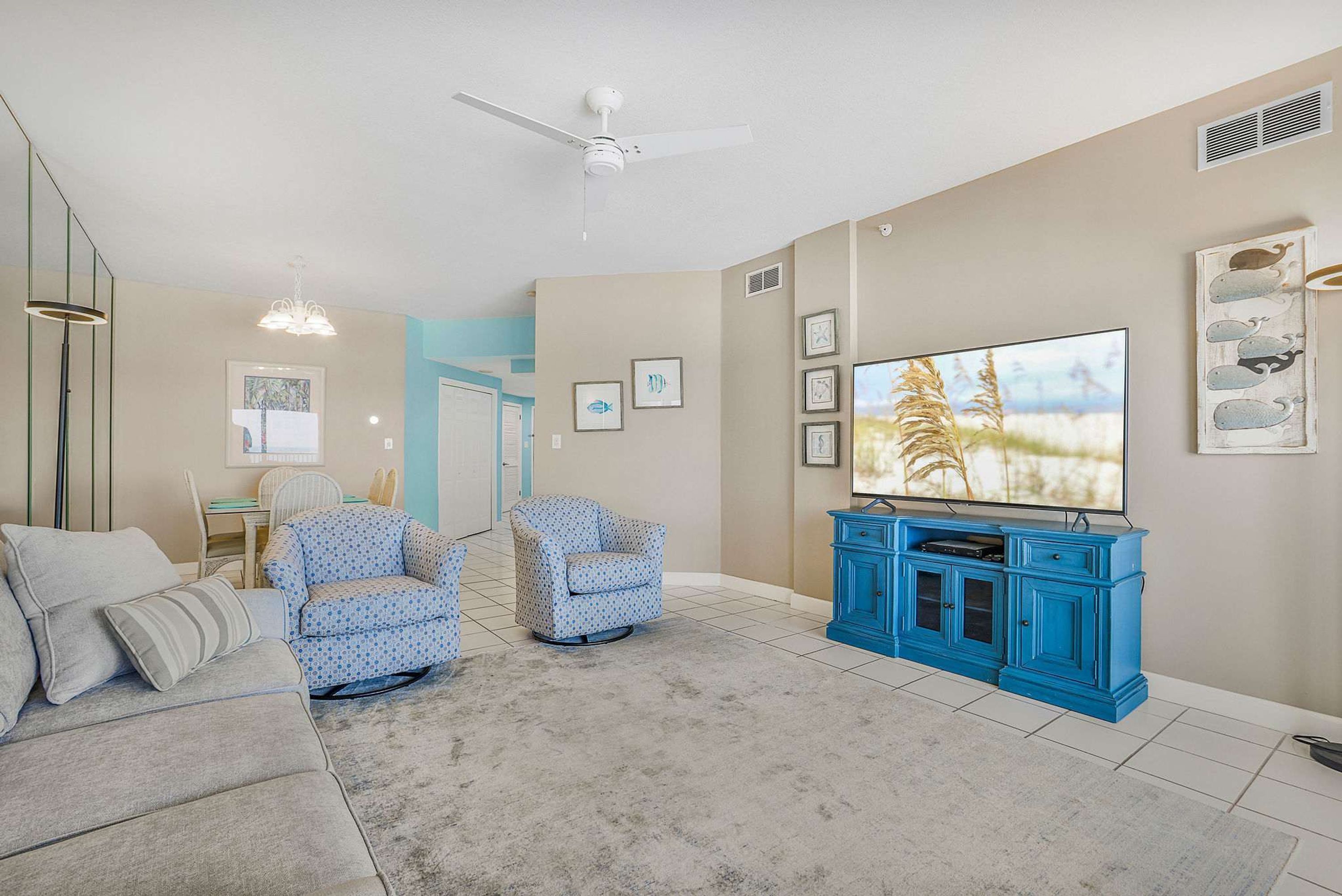 A living room with a grey sectional sofa, two patterned armchairs, a blue entertainment unit, and a ceiling fan.