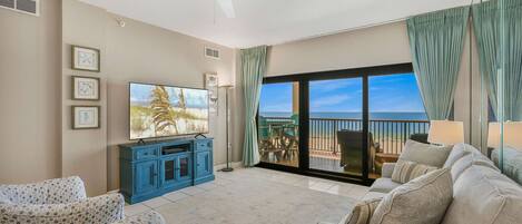 A living room with a large TV, blue cabinets, a beige sofa, and a balcony with a view.