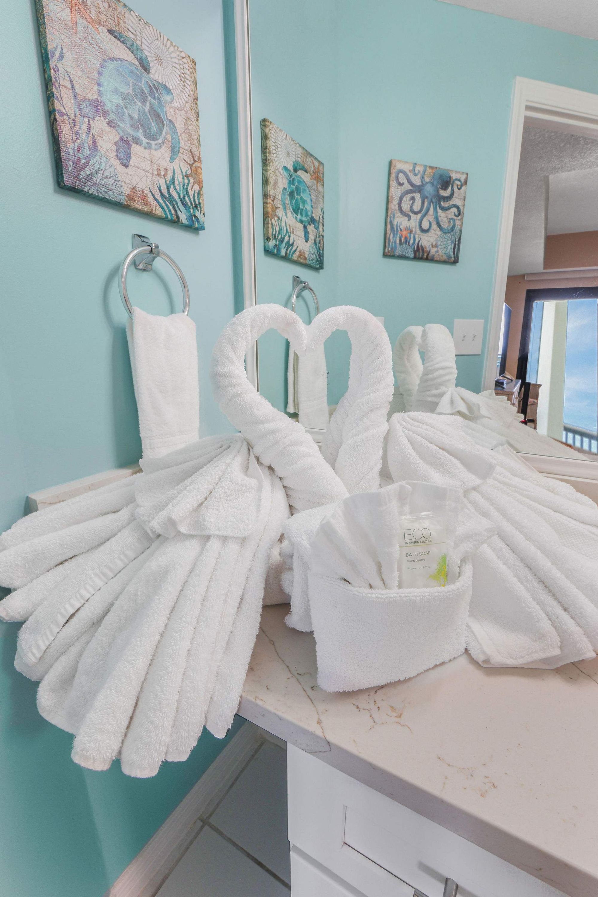 A bathroom with multiple white towels arranged on a counter, a glass container with a green object, and artwork on the walls.