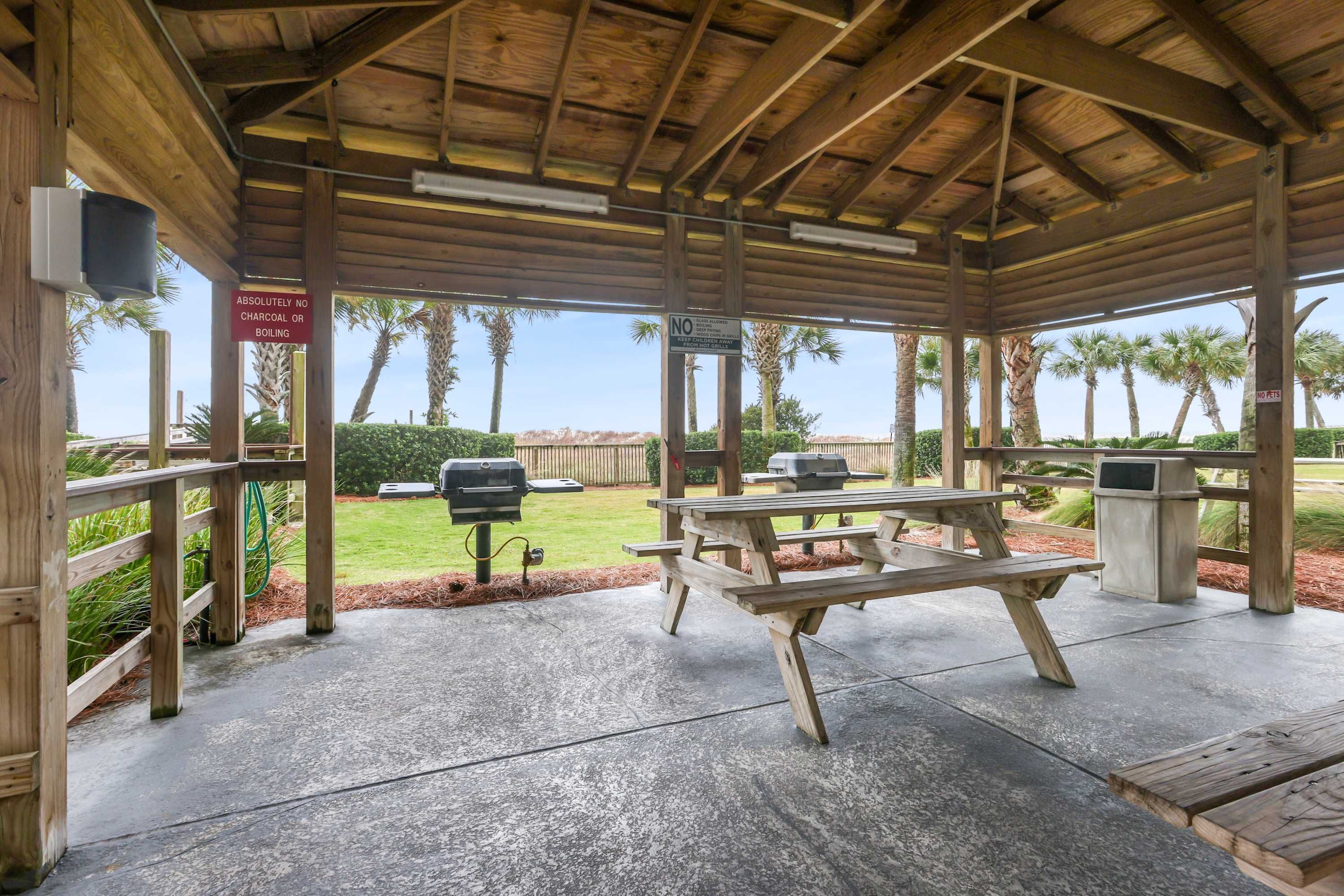 A covered outdoor area with picnic tables and barbecue grills, surrounded by palm trees and a grassy lawn.