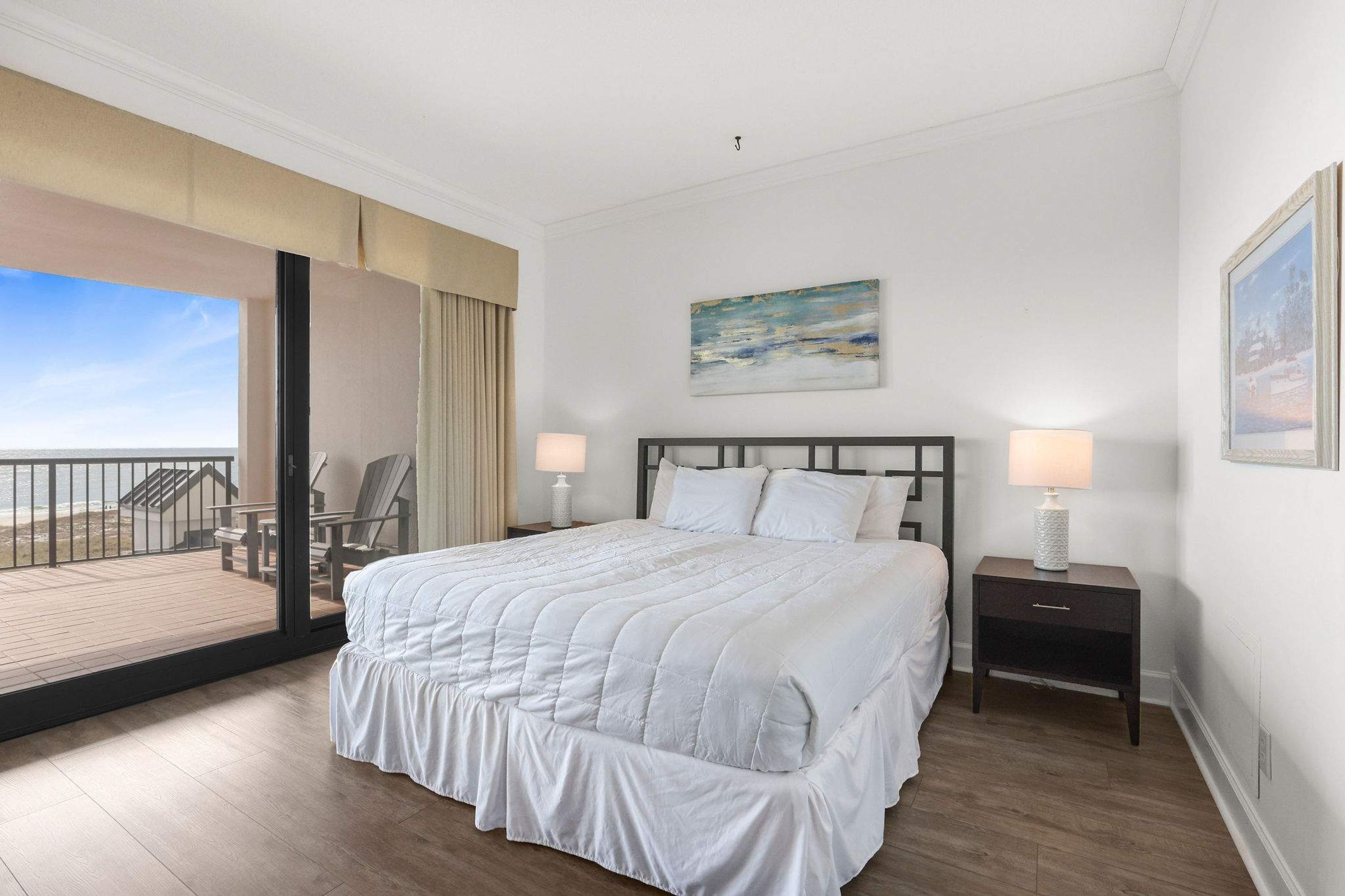 A bedroom with a large bed, a nightstand, a painting, and a view of the beach.