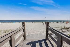 Ein hölzerner Steg führt zu einem Sandstrand bei strahlendem Himmel.