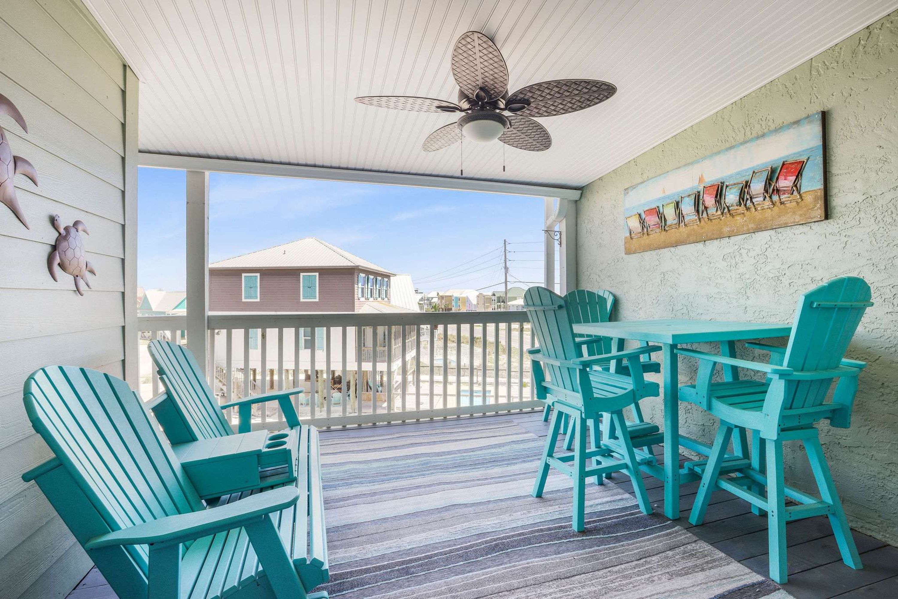 Un espace extérieur abrité, avec des fauteuils Adirondack de couleur turquoise et une table, un ventilateur de plafond et une peinture représentant des chaises de plage sur le mur.