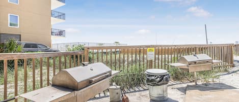 A barbecue setup with a grill and a trash can, located in a sandy area with a wooden fence and a building in the background.