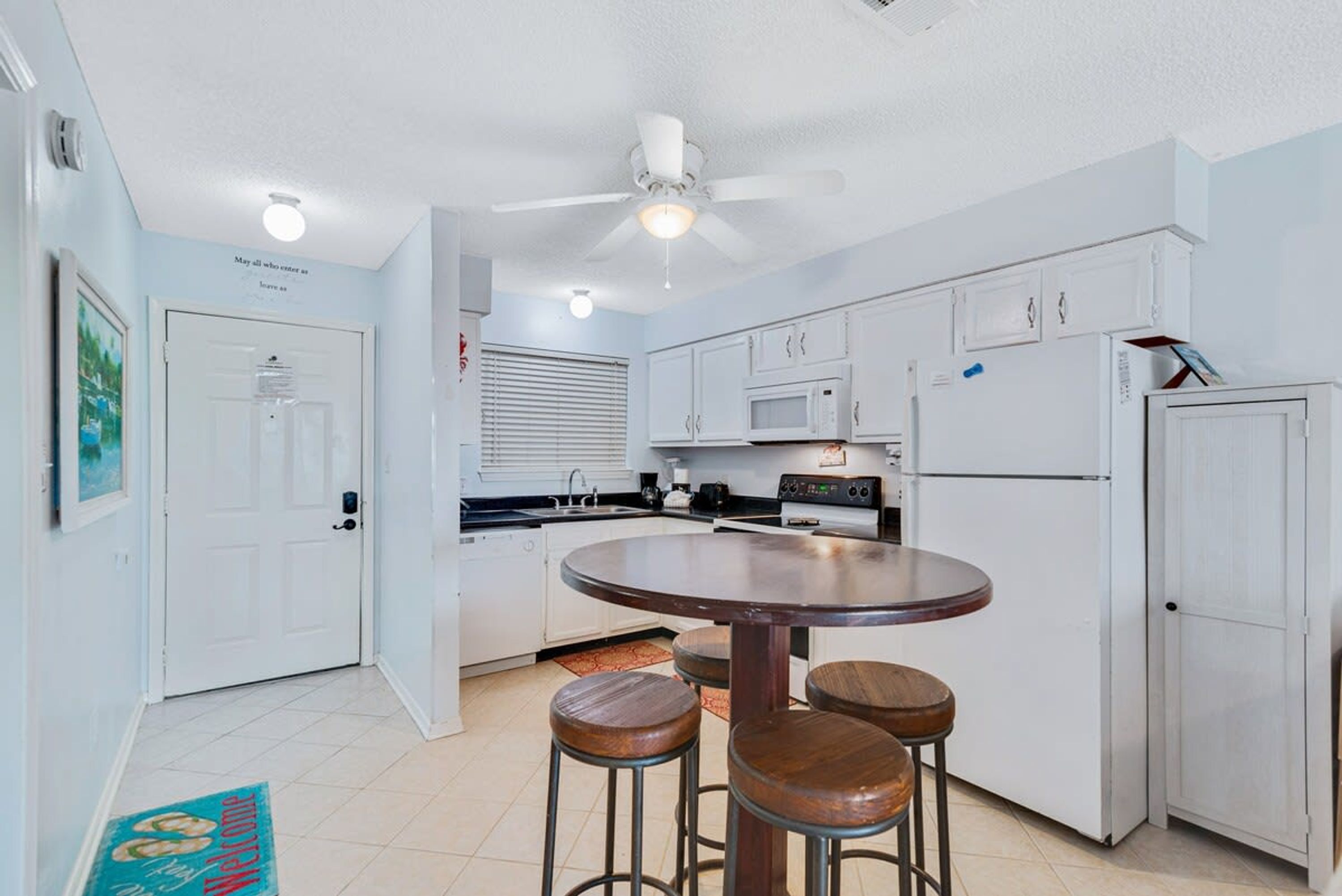 A compact kitchen with white cabinets, a microwave, a refrigerator, and a ceiling fan. There are three stools around a small round table.