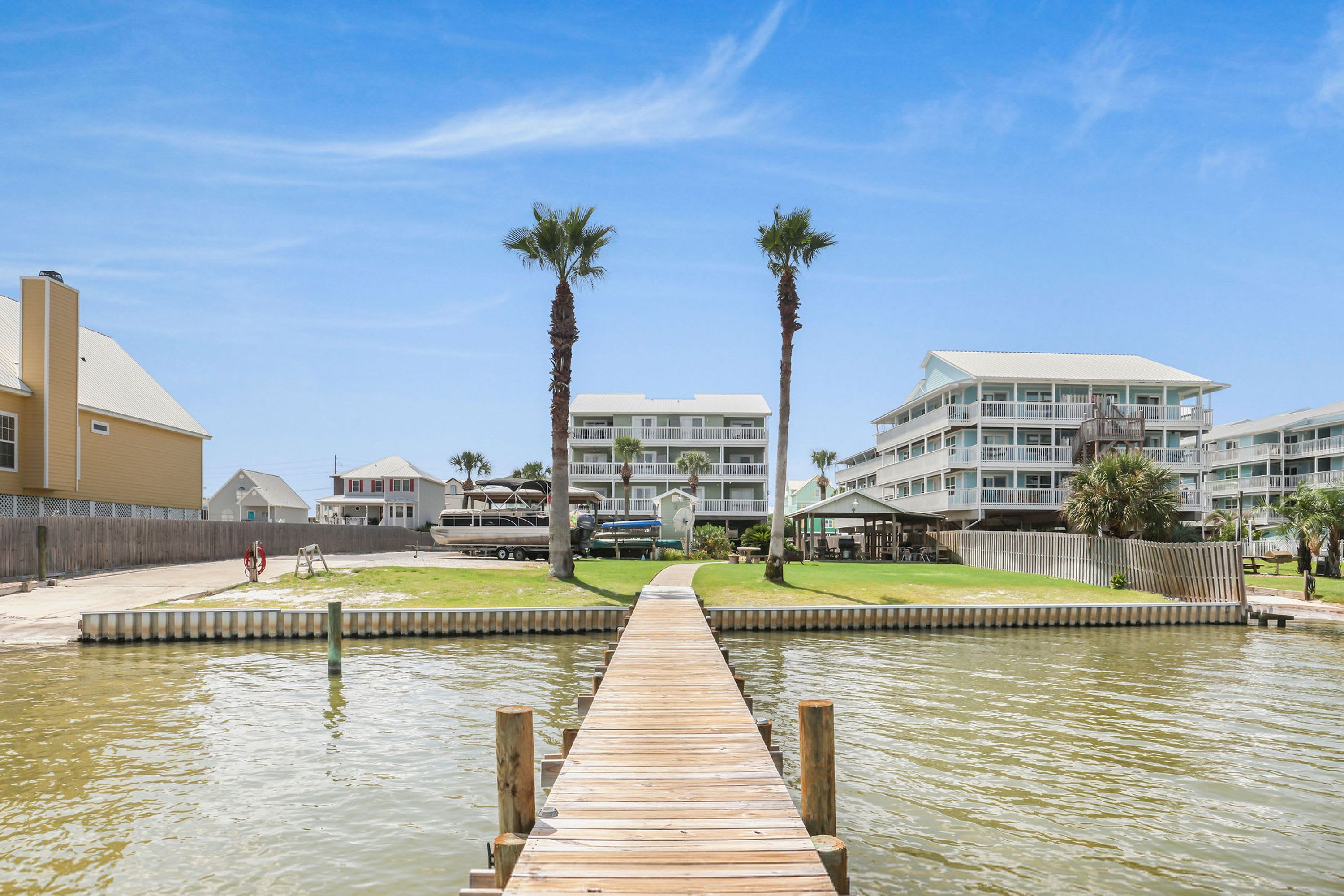A wooden pier leading to a waterfront area with palm trees and a building with multiple balconies.