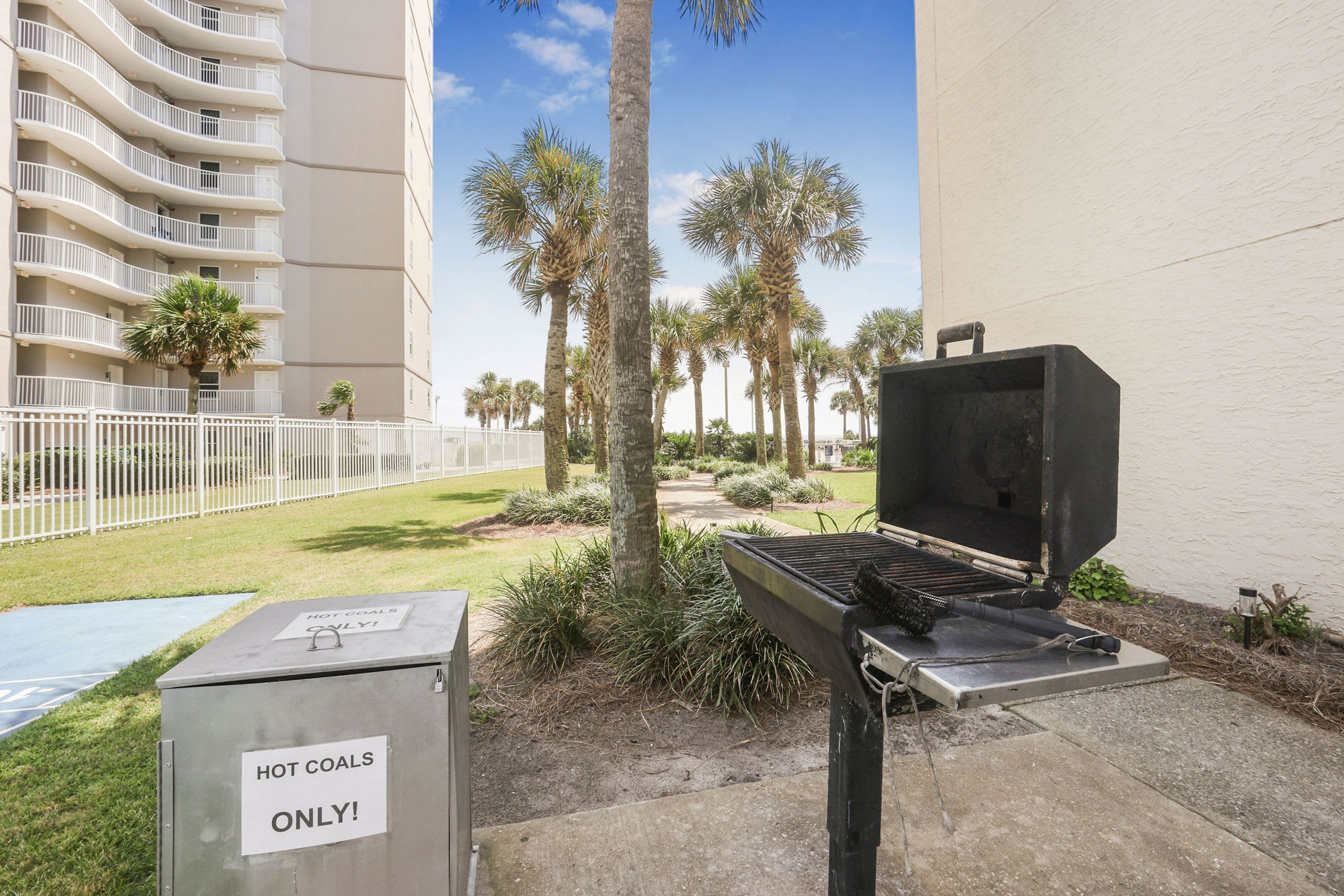 A barbecue grill with a sign indicating it's for hot coals only, surrounded by palm trees and a fenced area with a building in the background.