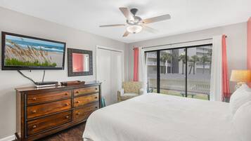 A hotel room with a bed, a dresser, a television, a ceiling fan, and a large window with a view of palm trees.