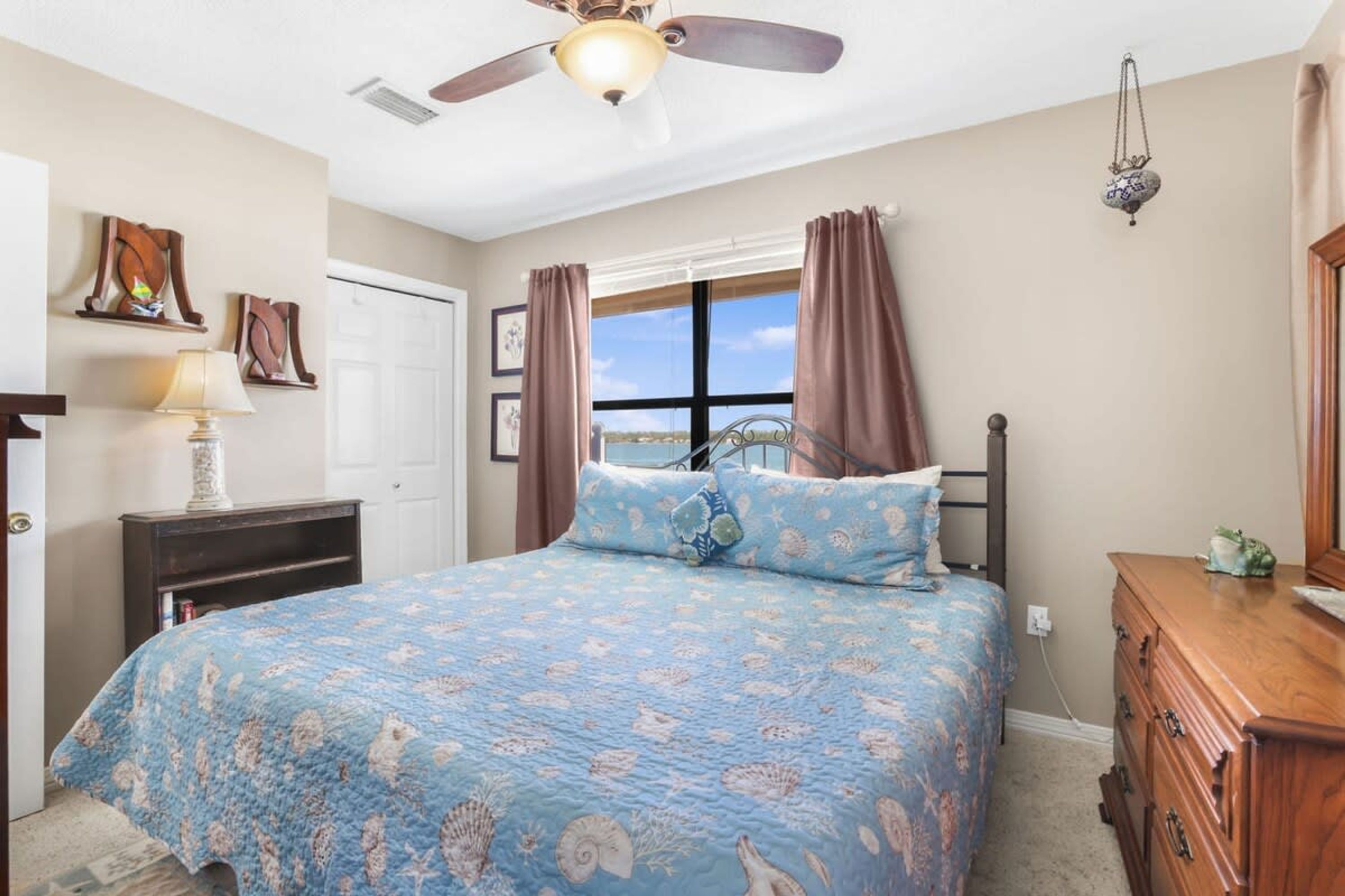Un dormitorio con cama, cómoda, ventilador de techo, ventana con cortinas y decoración en la pared.