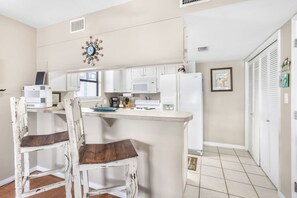 Cozinha compacta com armários brancos, micro-ondas e geladeira. Duas cadeiras de madeira com almofadas escuras. Um relógio decorativo na parede e uma pequena janela que traz luz natural.