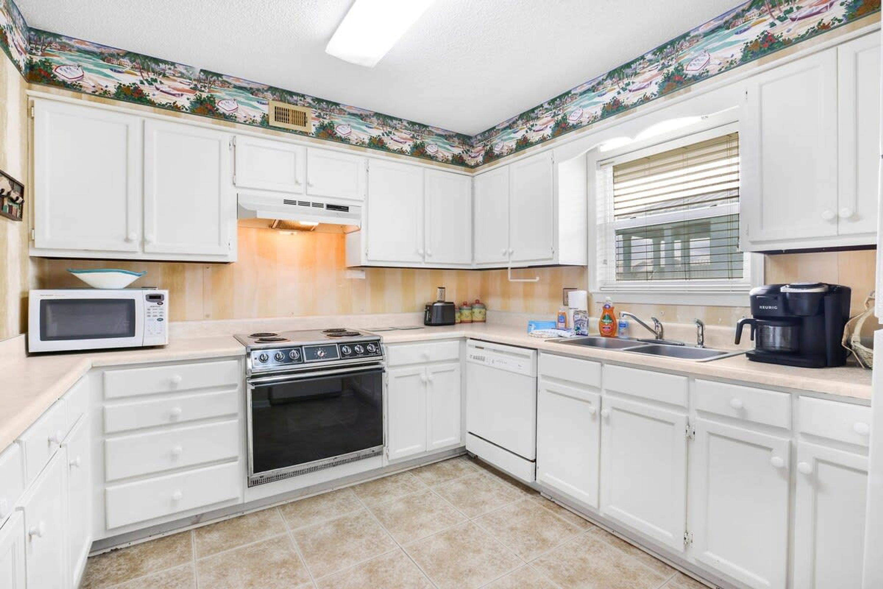 Una cocina con armarios blancos, microondas, estufa, horno, lavavajillas y una ventana con persianas.