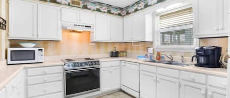 Una cocina con armarios blancos, microondas, estufa, horno, lavavajillas y una ventana con persianas.