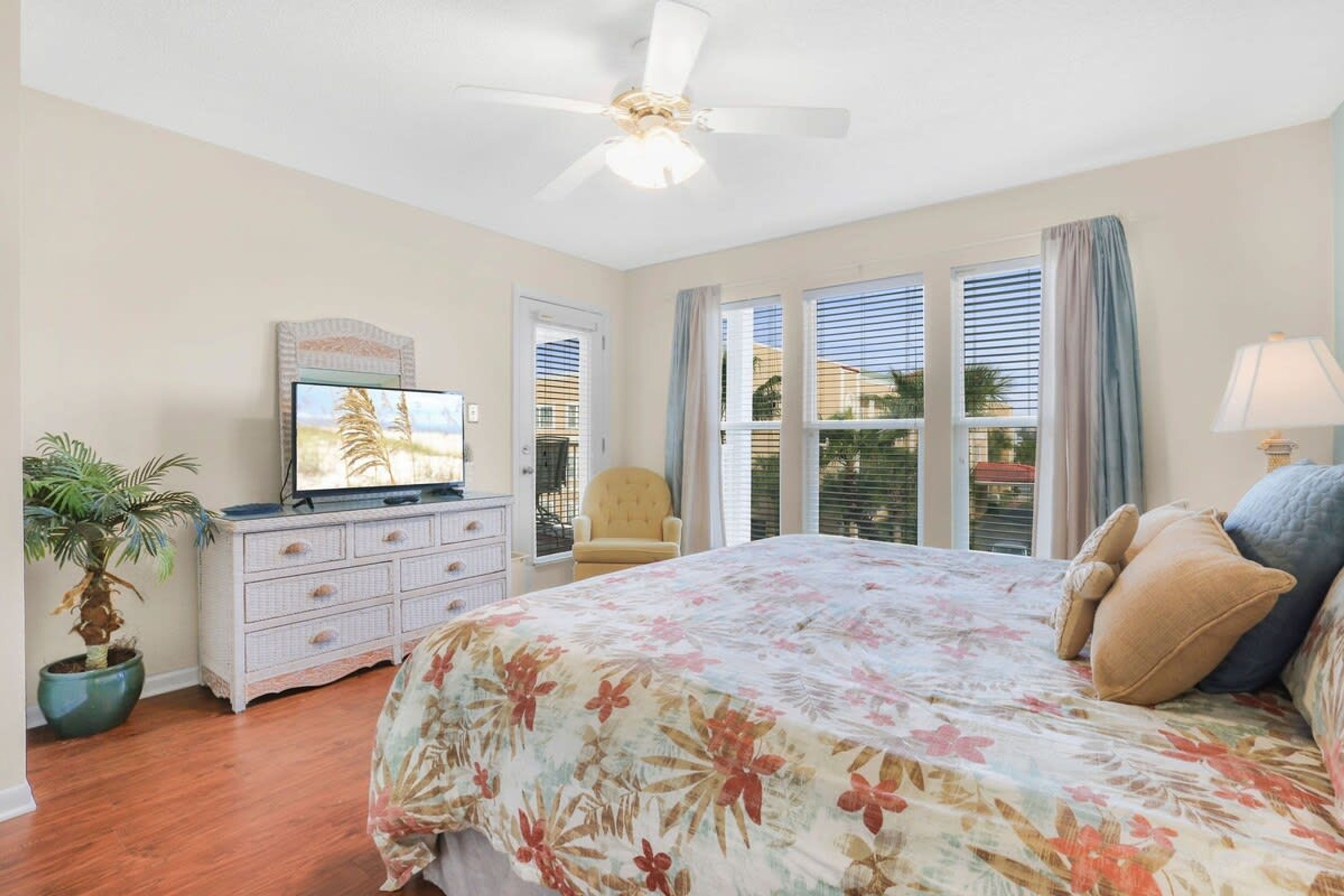Un dormitorio con cama, televisión, ventilador de techo, cómoda, silla y ventana con persianas.