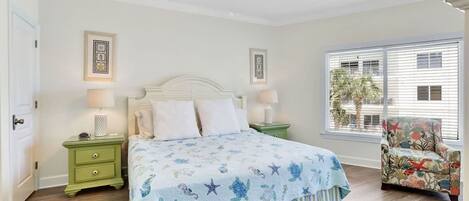 Une chambre à coucher comprenant un lit, deux tables de chevet, une chaise, un ventilateur de plafond et une fenêtre avec des stores.