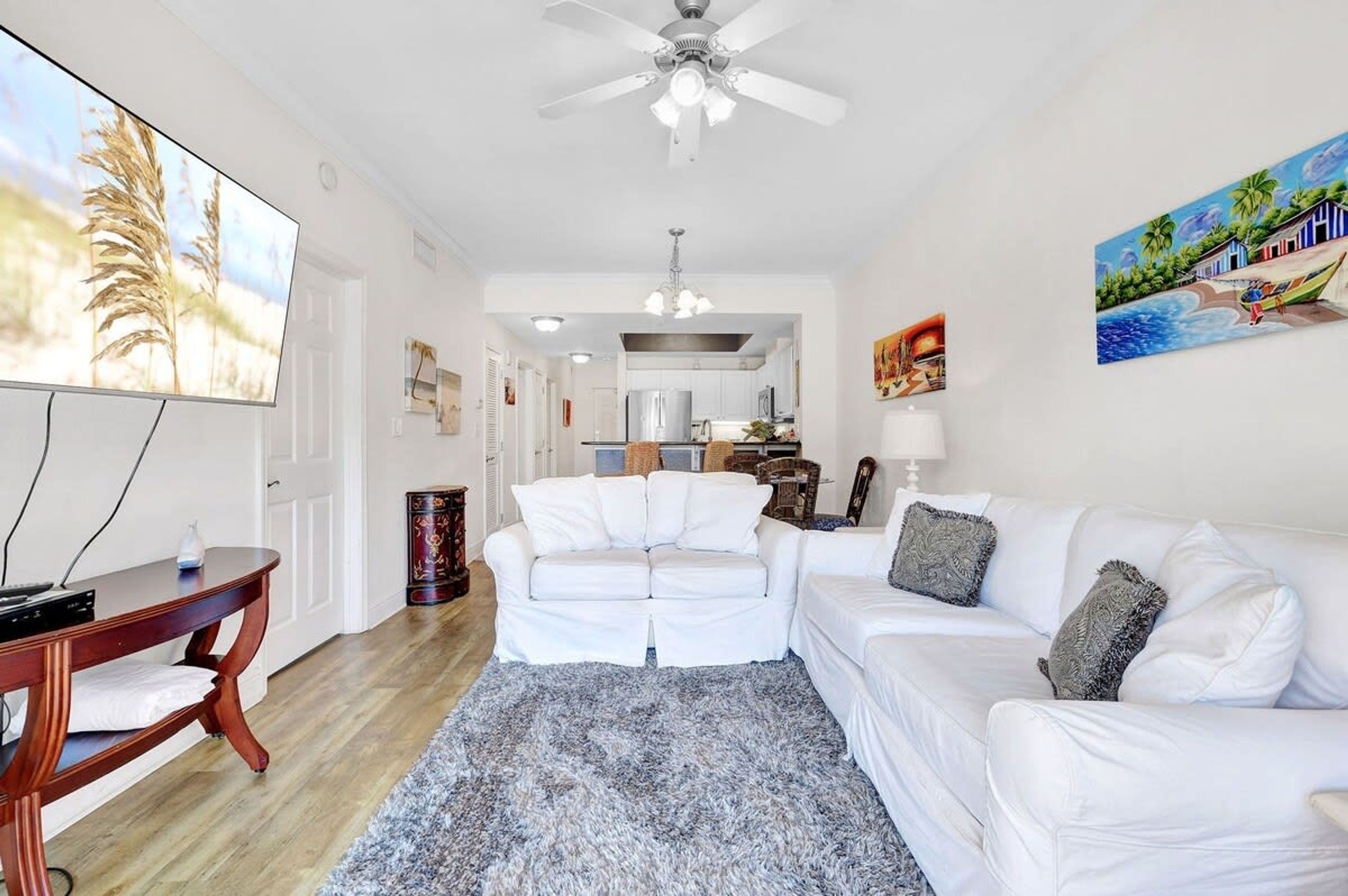 A living room with a white sofa, a coffee table, and a TV. There is a ceiling fan, a rug, and a painting on the wall.