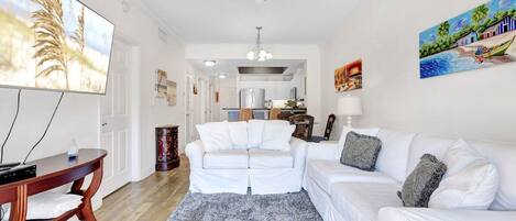 A living room with a white sofa, a coffee table, and a TV. There is a ceiling fan, a rug, and a painting on the wall.