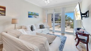 Een moderne woonkamer met een witte zithoek, een plafondventilator en een groot raam met uitzicht op een balkon.