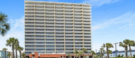A multi-story hotel with a uniform facade, a parking lot with several cars, and palm trees around.