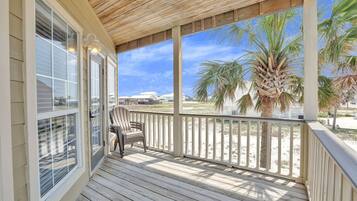 A wooden deck with a chair, overlooking a beach with palm trees and houses in the background.