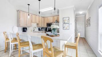 A modern kitchen with light wooden cabinets, a central island with bar stools, pendant lighting, and a view into the adjacent dining area.