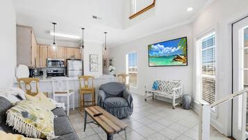 A modern living room with a sofa, a coffee table, and a TV. There is a kitchen area with wooden cabinets and a dining table.