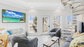 A modern living room with a large TV, comfortable seating, and a view of the ocean through large windows.
