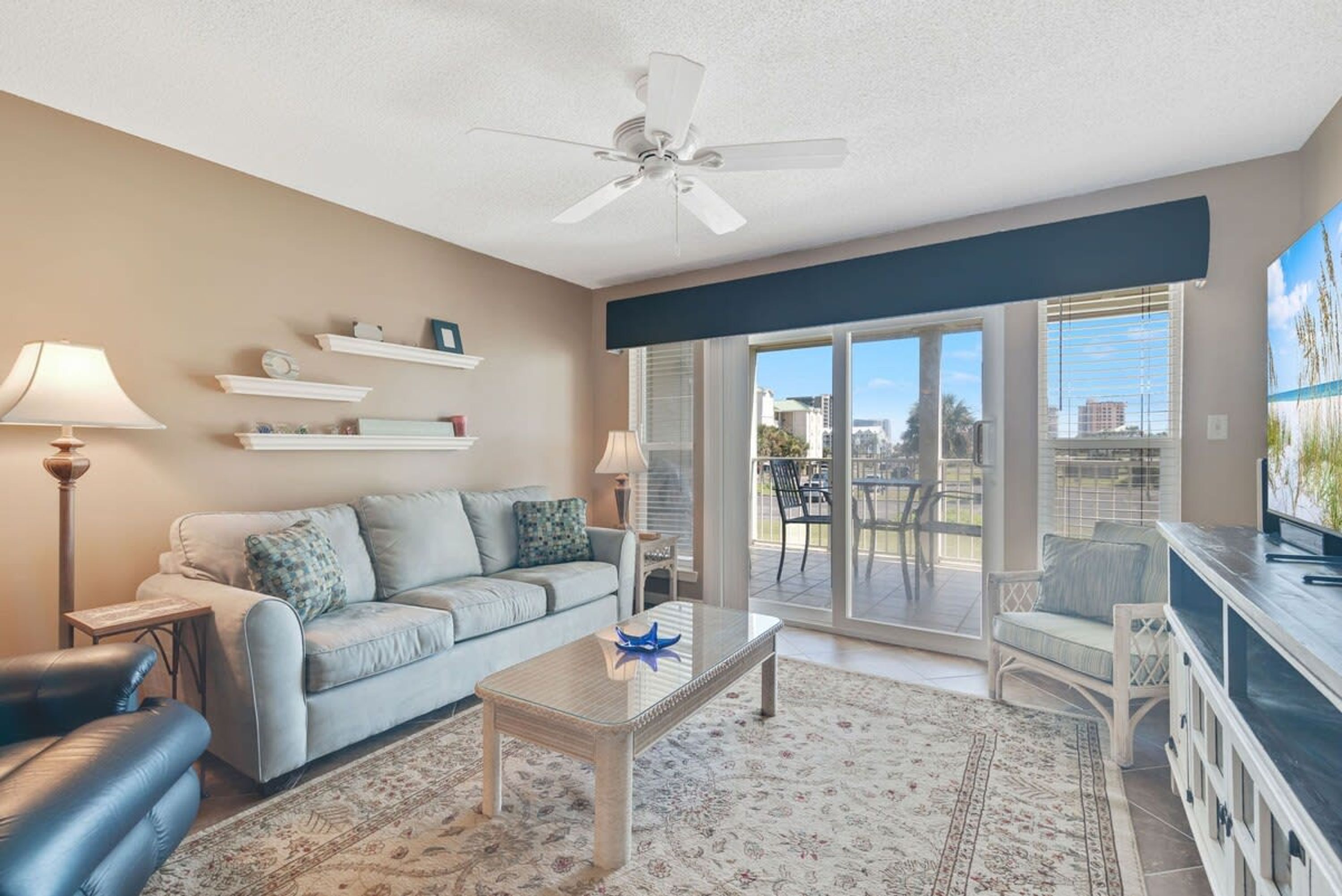 A living room with a sofa, coffee table, and a ceiling fan. There are shelves with decorative items and a view of the outdoors through a sliding glass door.