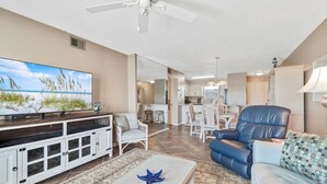 A spacious living room with a flat-screen TV, a ceiling fan, and a dining area with a kitchen in the background. - Portside 108- On Ole River Bring Boat Pool (Orange Beach)