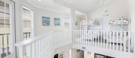 A spacious, well-lit room with a high ceiling, a white staircase, a kitchen area, and bunk beds with nautical-themed bedding.